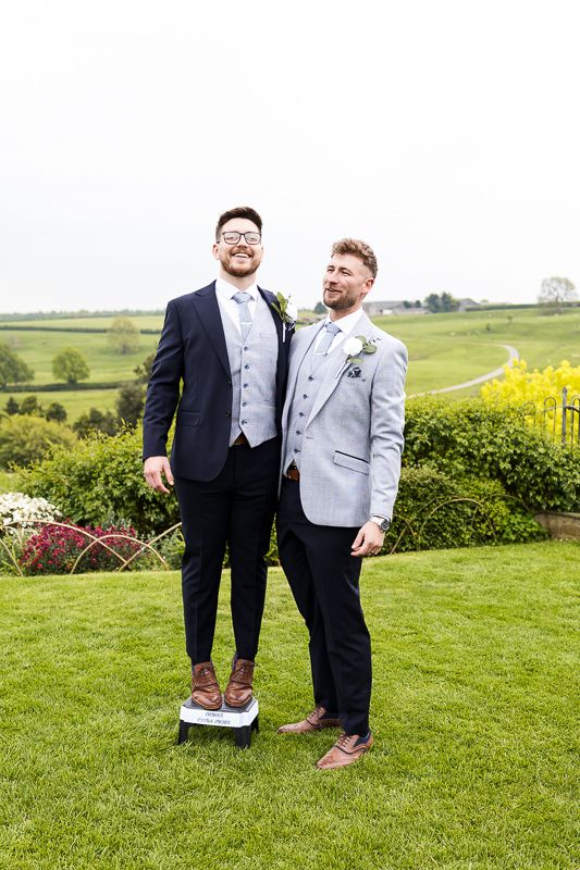 Two men in formal wedding attire standing together on a manicured lawn with countryside views in the background.