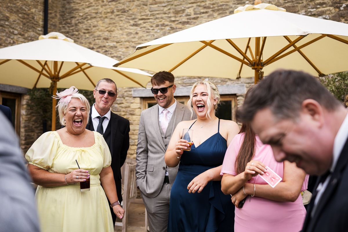 Wedding guests socializing outdoors under cream umbrellas at a reception venue with stone wall backdrop.