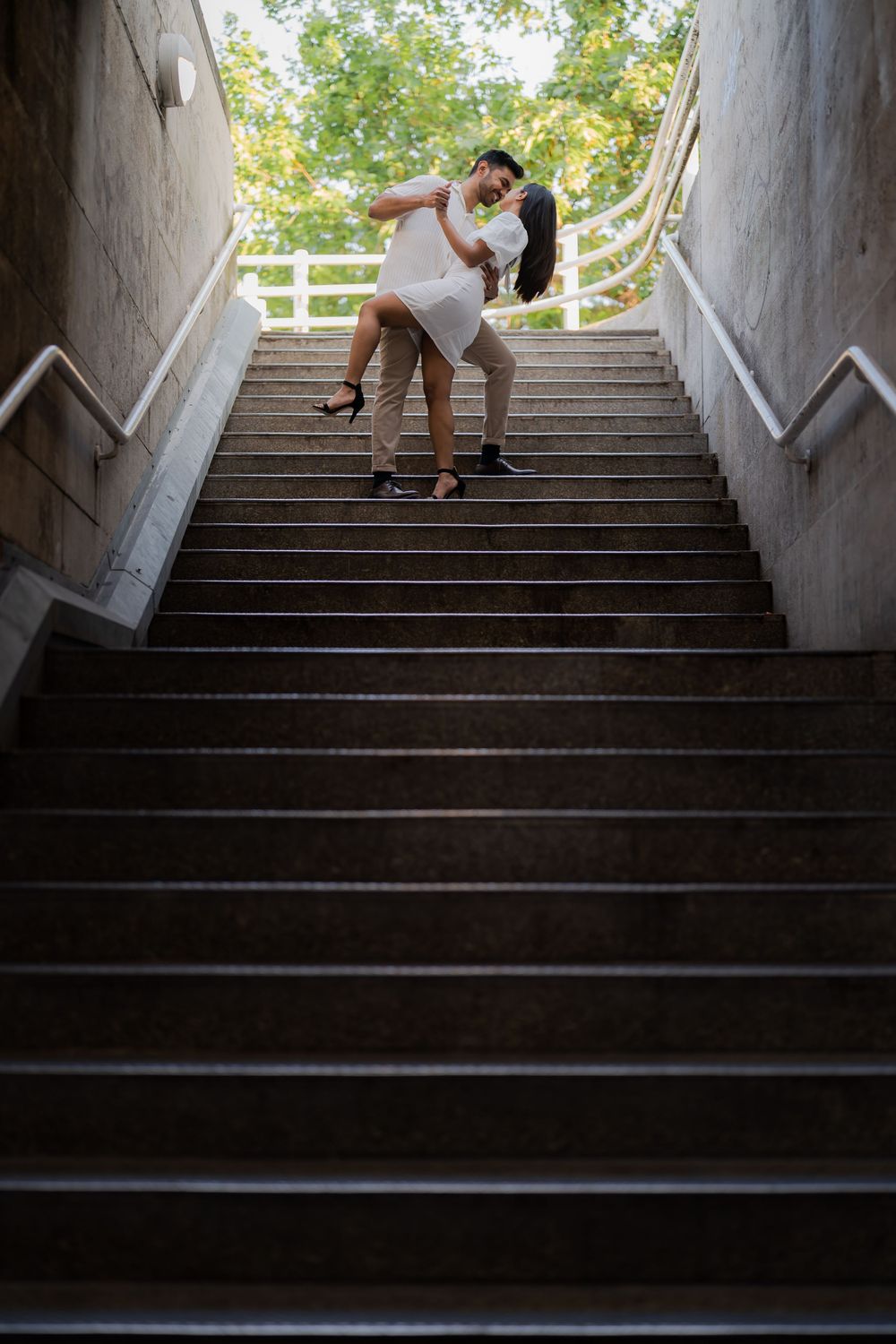 Couple embracing romantically on outdoor stone staircase, woman in white dress and heels, man in casual attire, lush trees above.
