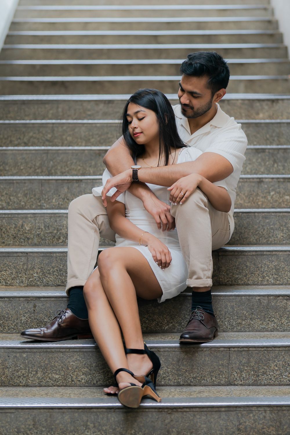 A couple sits intimately on stone steps, woman in white dress with heels, man in beige outfit with watch embracing her.