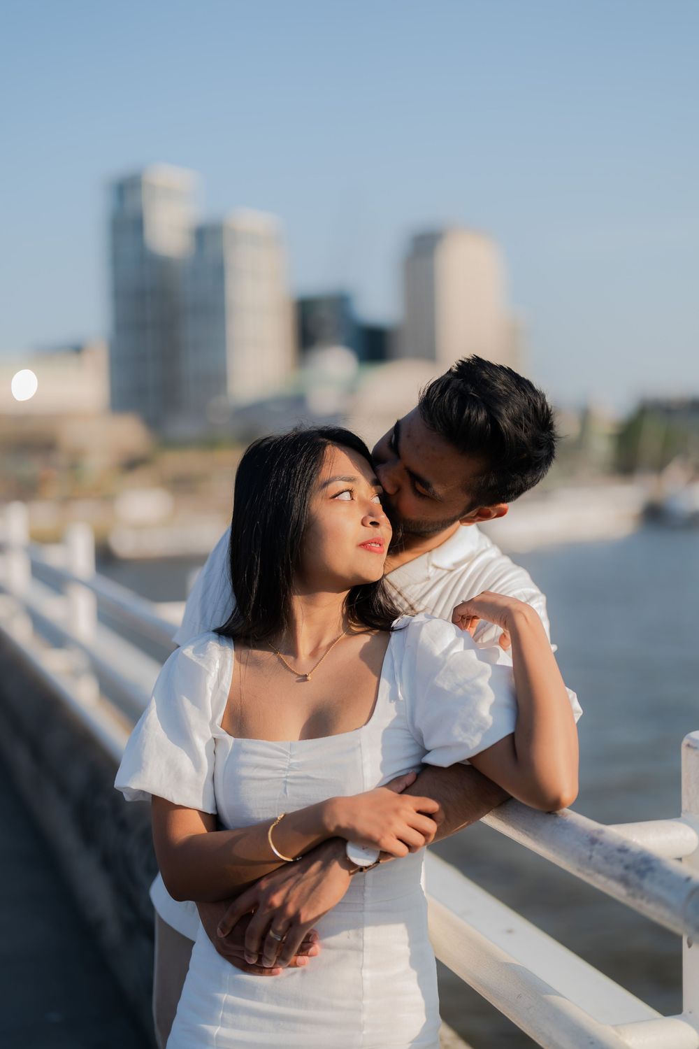 Couple embracing on a waterfront bridge with city skyline, man kissing woman's cheek in matching white outfits.