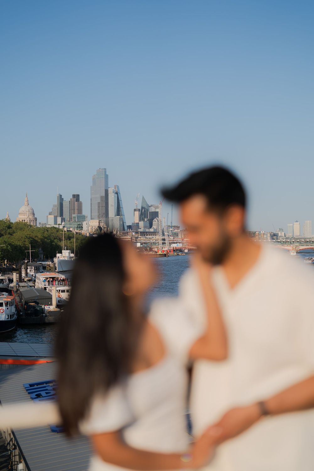 Couple in white embracing on a bridge with London skyline and River Thames in the background on a clear day.