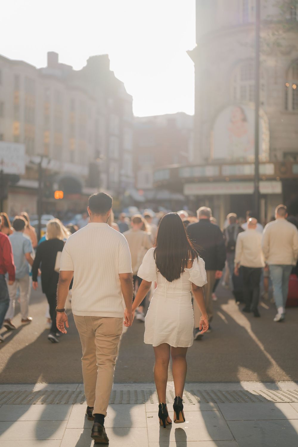 Couple holding hands walking through a busy sunlit city street, viewed from behind, dressed in white and beige outfits.