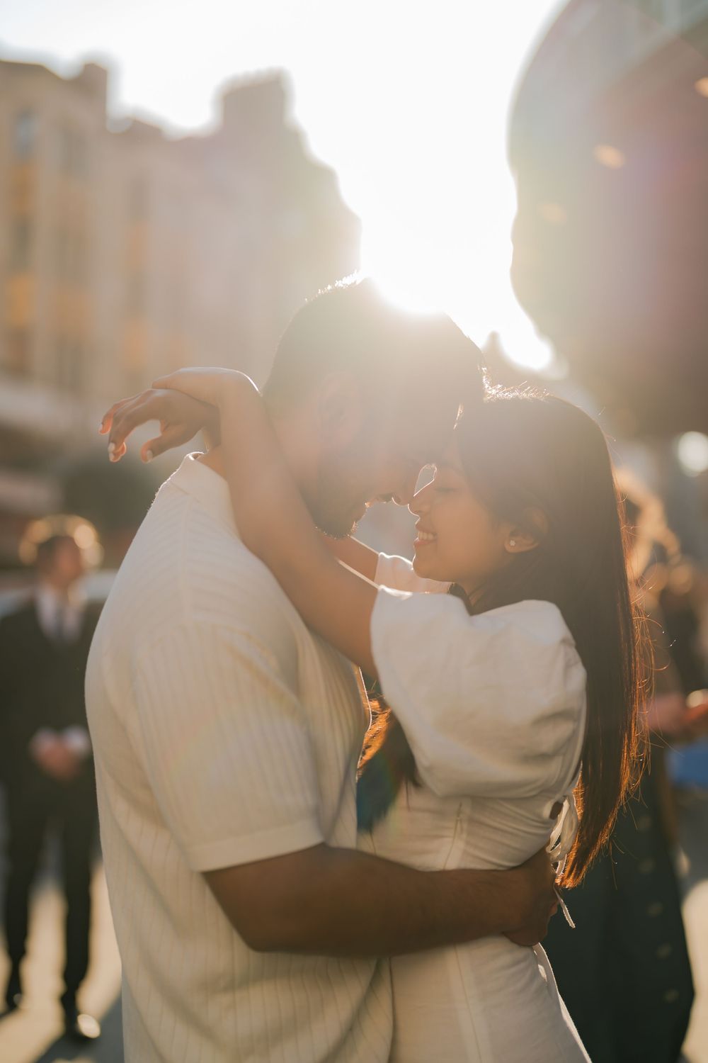 Couple embracing and almost kissing in golden sunlight, backlit by warm sun flare in an outdoor urban setting.