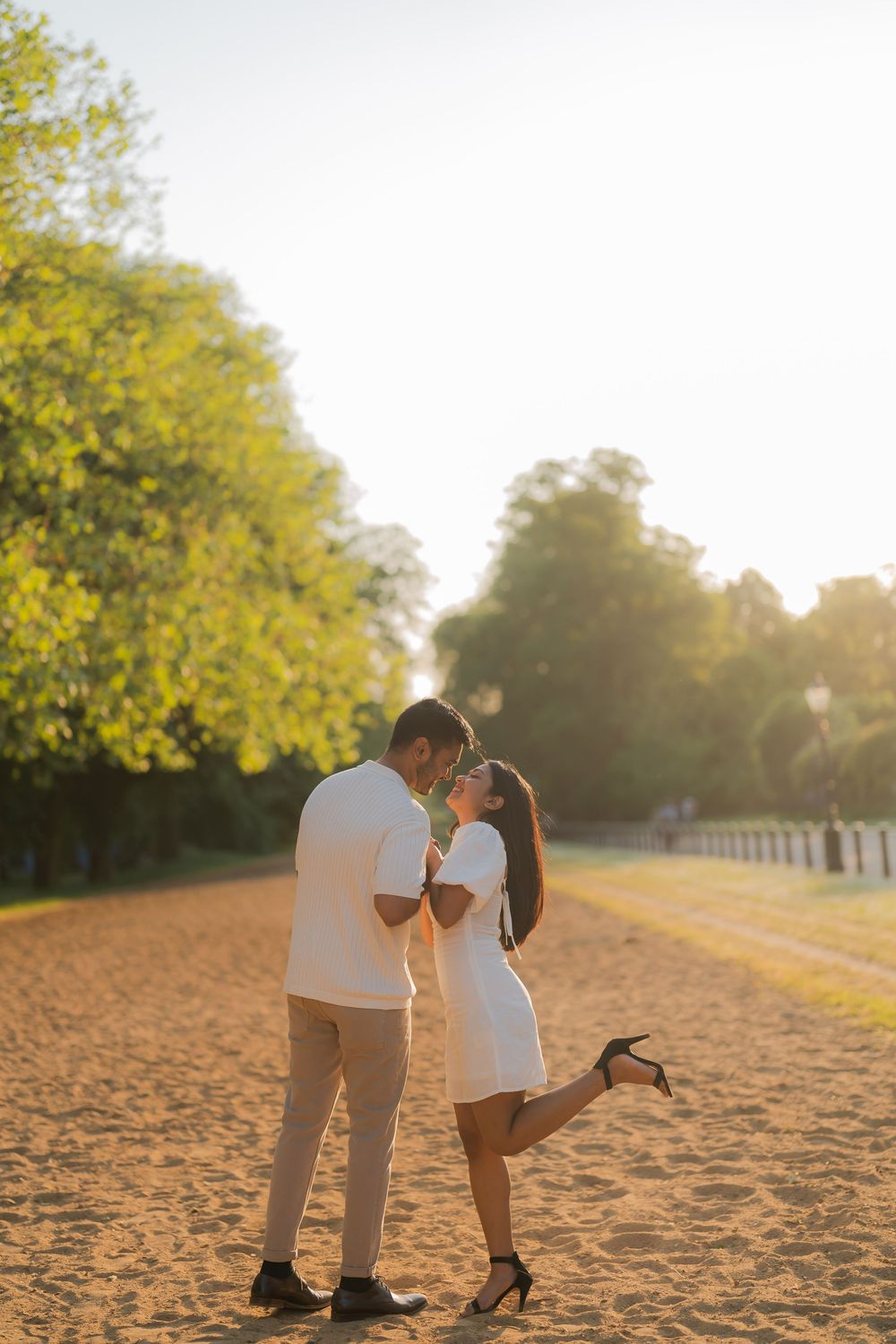 Romantic couple in white outfits sharing a kiss at golden hour on a sandy path surrounded by lush green trees.