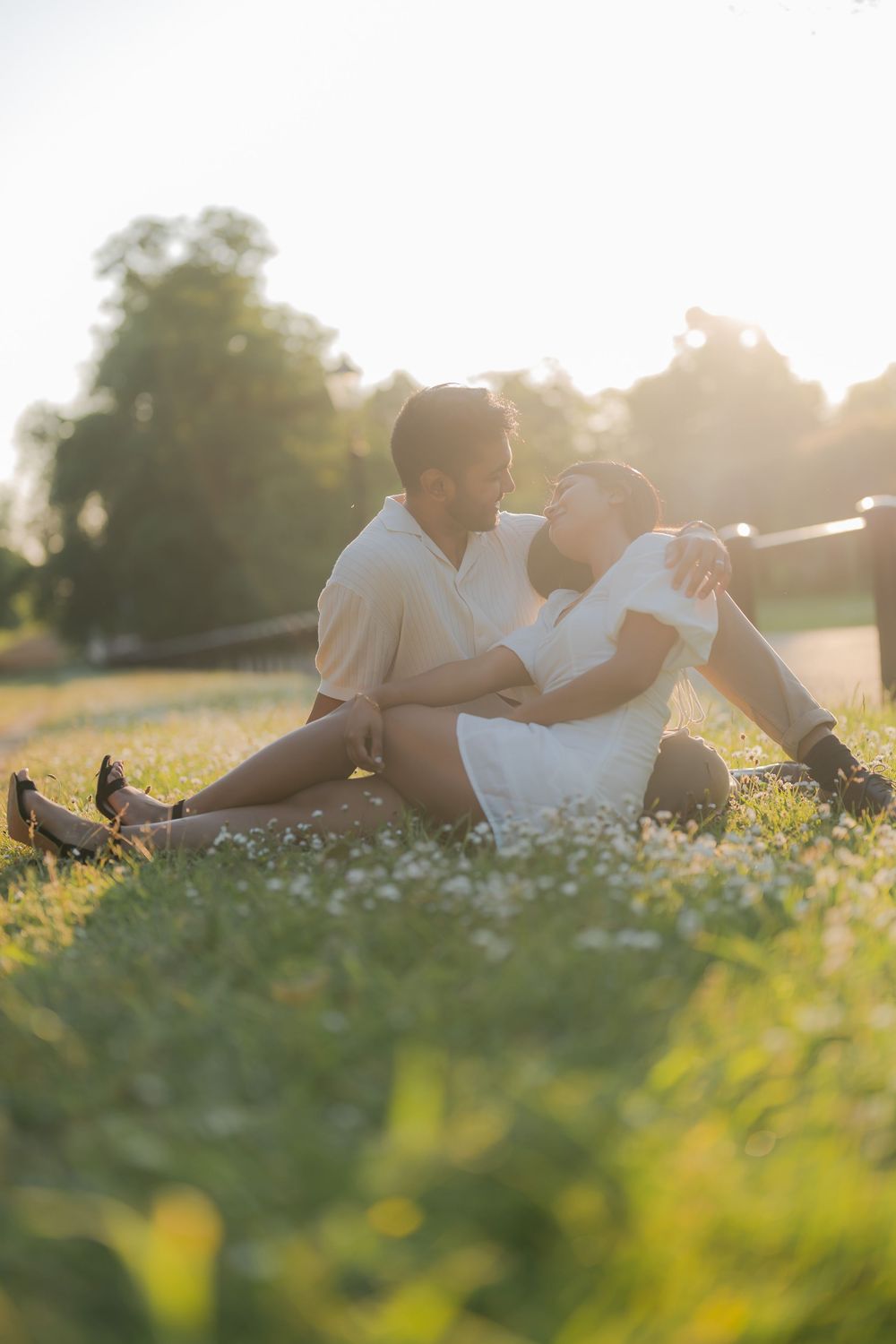 Couple sitting together on a sunlit grassy field with wildflowers, sharing a romantic moment at golden hour.