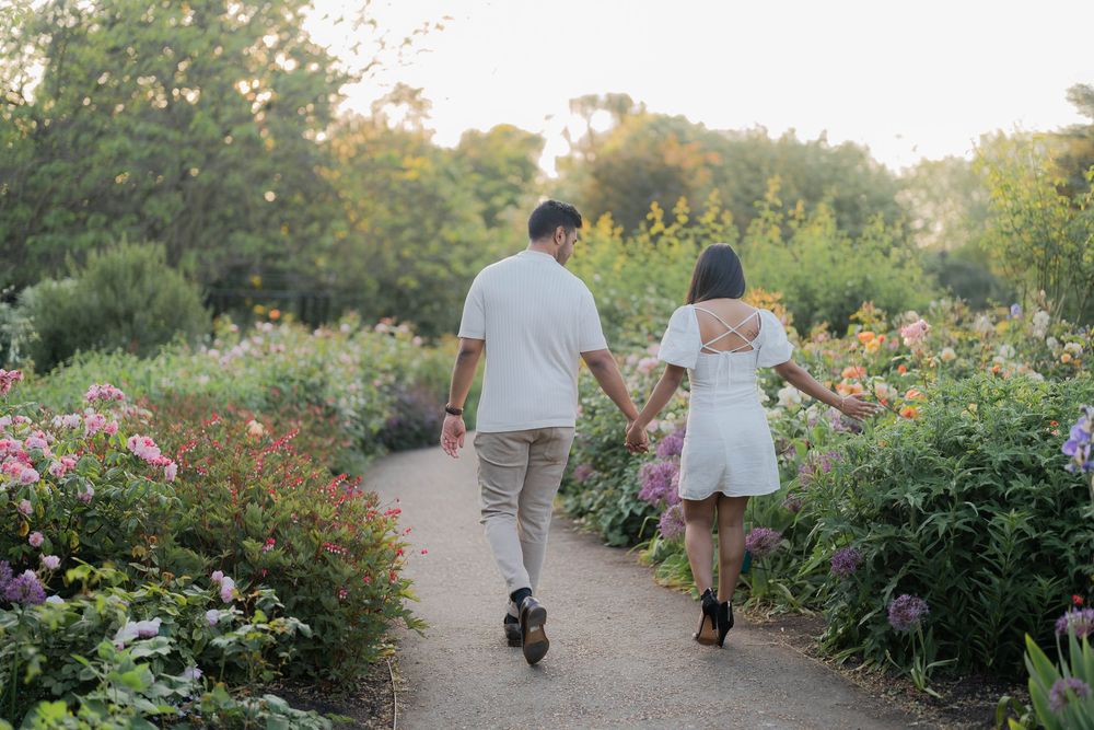 A couple holds hands walking down a garden path surrounded by colorful blooming flowers at sunset.
