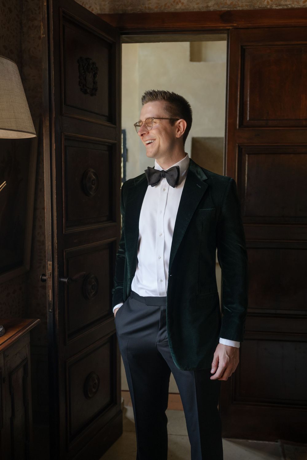 A smiling man in a green velvet tuxedo jacket, bow tie, and glasses stands confidently in an elegant wood-paneled room.