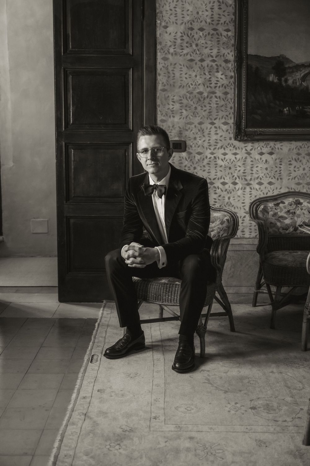 Elegant man in tuxedo and bow tie seated in vintage chair, black and white wedding portrait.