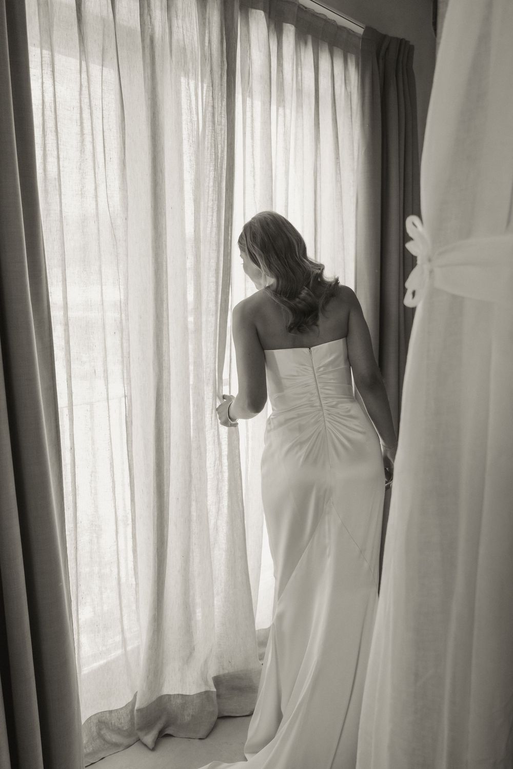Bride in white strapless wedding gown standing at window with sheer curtains, black and white photo.