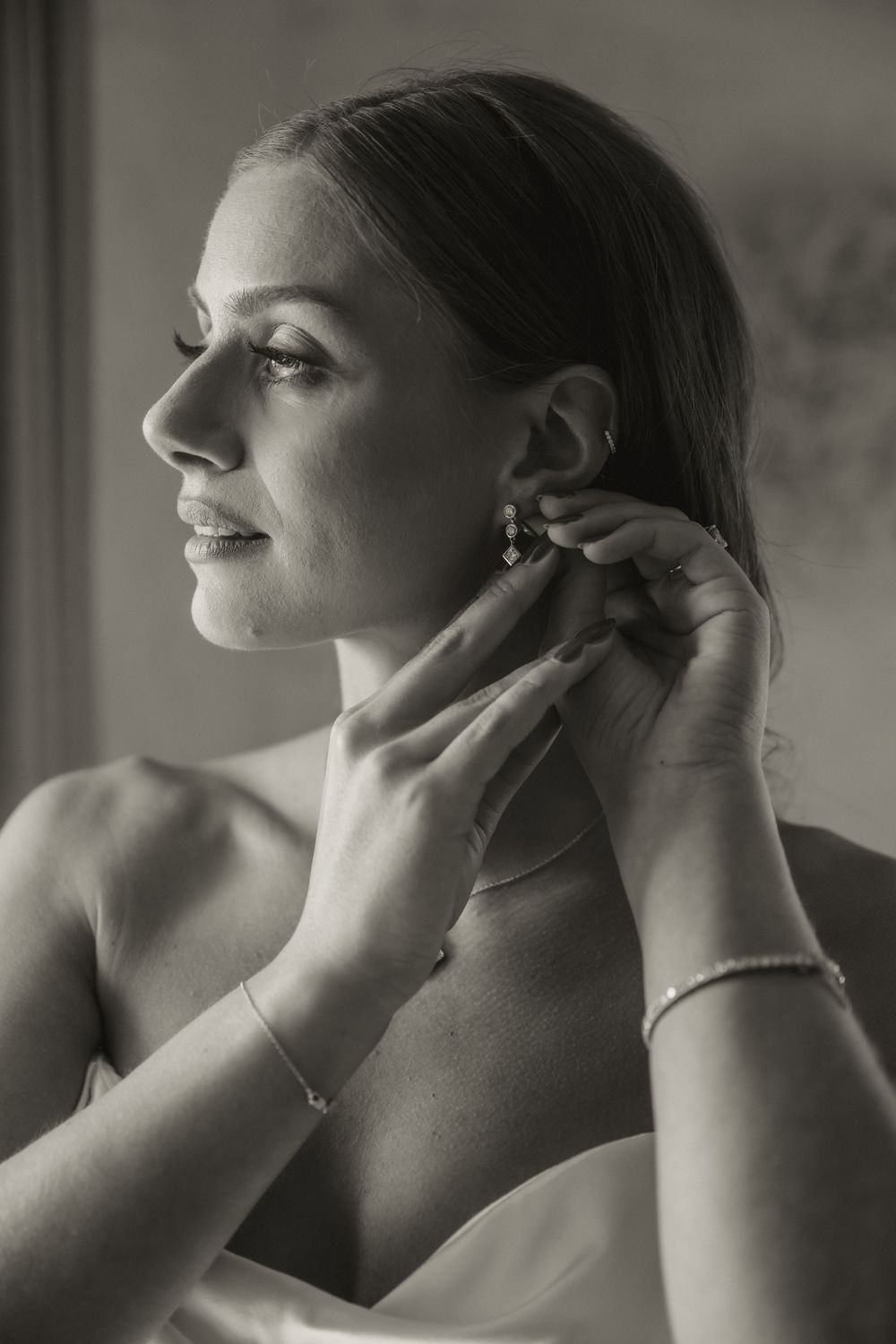 Bride putting on earrings in black and white wedding portrait, wearing a bracelet and strapless gown.