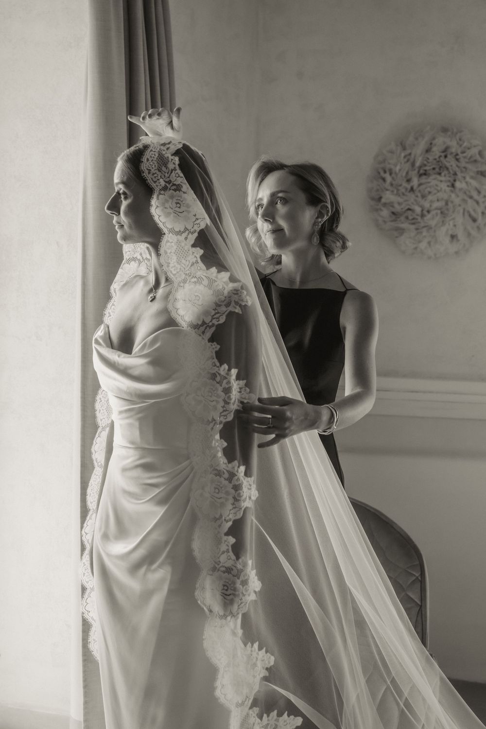 Bride in floral lace wedding gown with veil assisted by bridesmaid near window, black and white.