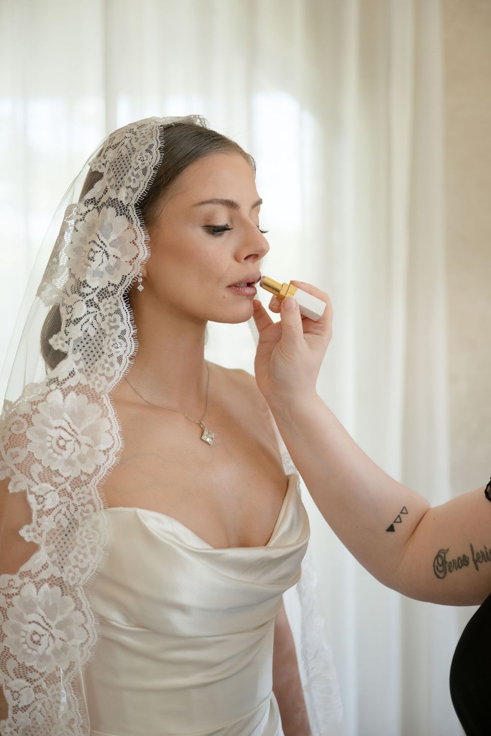 Bride in white satin gown and lace veil gets lipstick applied by makeup artist with tattooed arm.