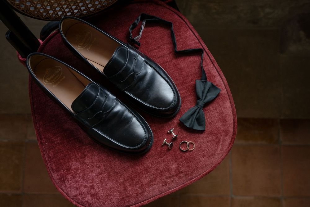 Black leather penny loafers with bow tie, cufflinks, and rings on a red velvet chair for a formal wedding ensemble.