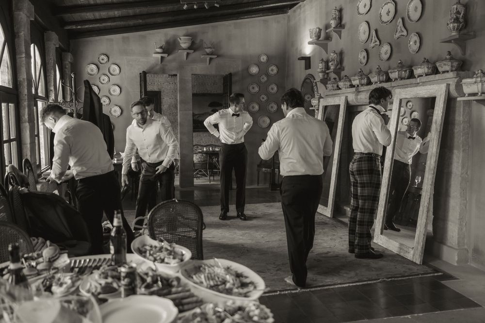Black and white photo of groomsmen preparing in elegant room with mirrors, decorated walls, and dining table.