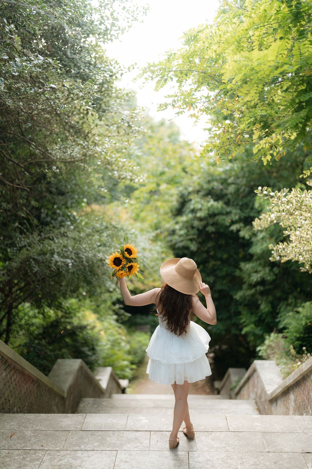 Woman in white dress and sun hat holding sunflowers on stone steps surrounded by lush green trees.