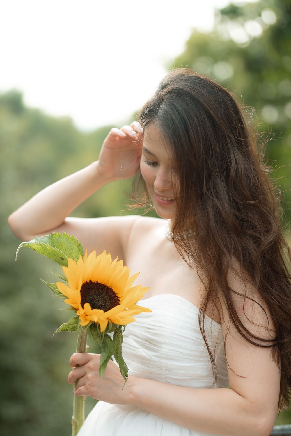 Young Asian woman in white strapless dress holding a sunflower outdoors, smiling softly with long dark hair flowing.