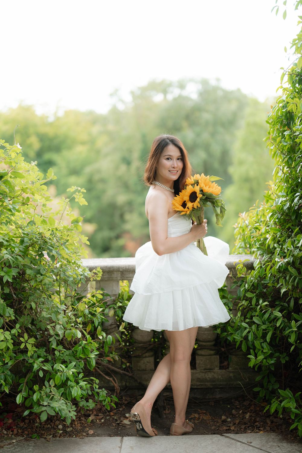 Young woman in white dress holding sunflowers bouquet, smiling outdoors near stone balustrade surrounded by lush greenery.