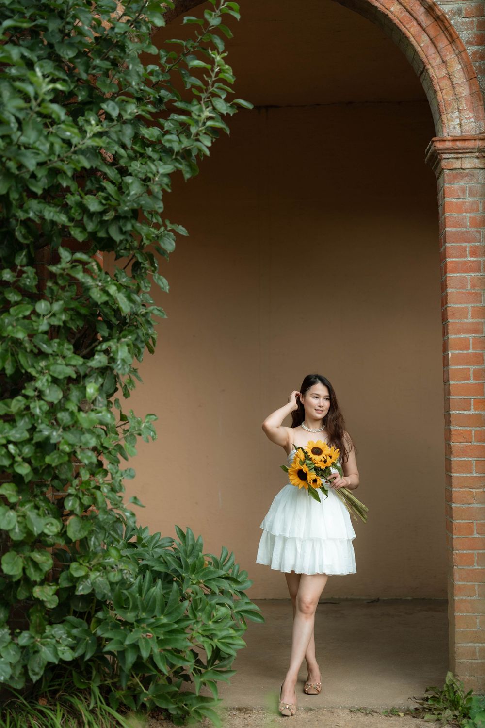 Young Asian woman in white dress holding sunflowers stands under brick archway surrounded by lush green ivy.