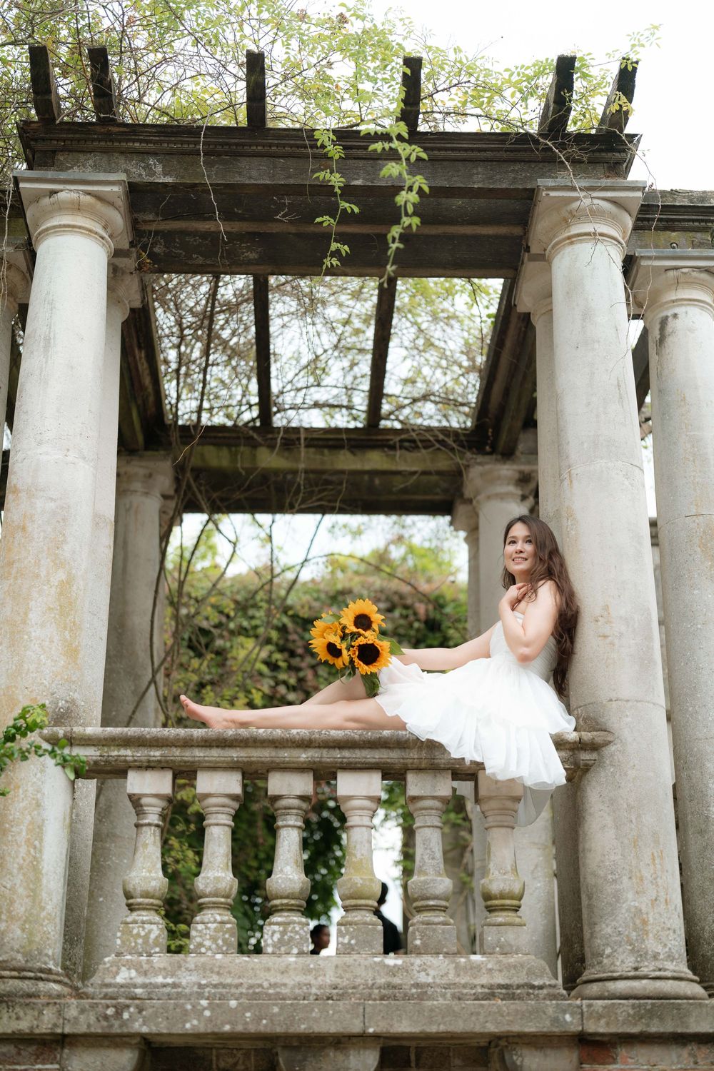 Ballerina in white tutu sits on stone balustrade holding sunflowers at a classic garden pavilion with columns.