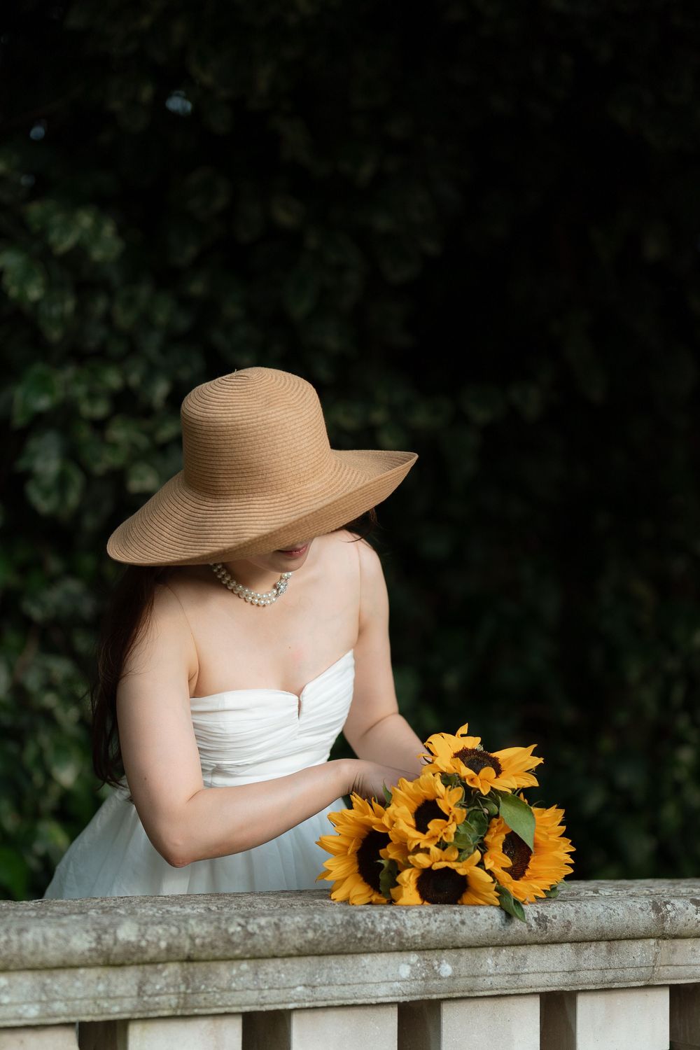 Woman in white strapless dress and wide-brim straw hat holding sunflowers by a stone balustrade outdoors.