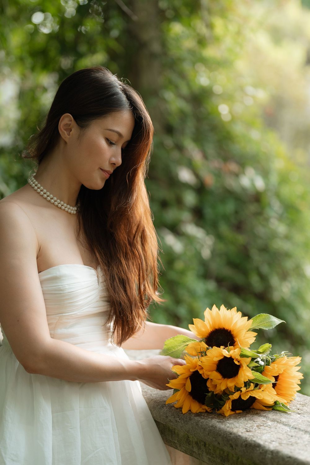 A young Asian bride in a white strapless gown holds a sunflower bouquet outdoors, wearing a pearl necklace.