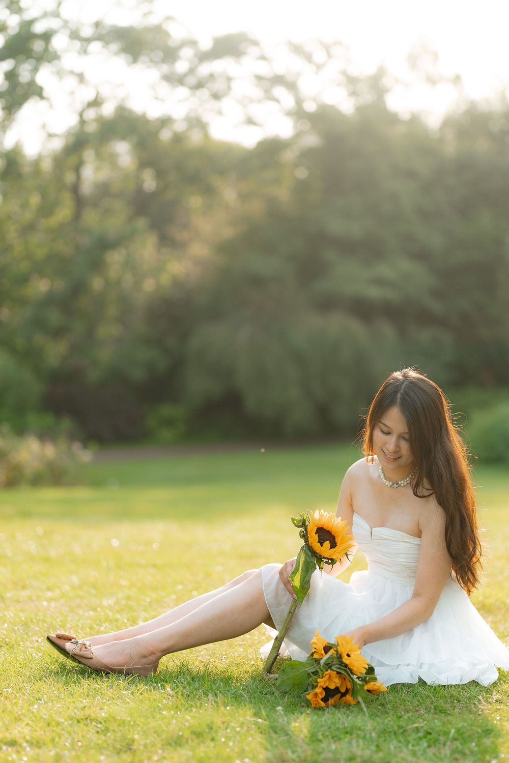Young Asian woman in white strapless dress sitting on grass holding sunflowers in golden hour sunlight.