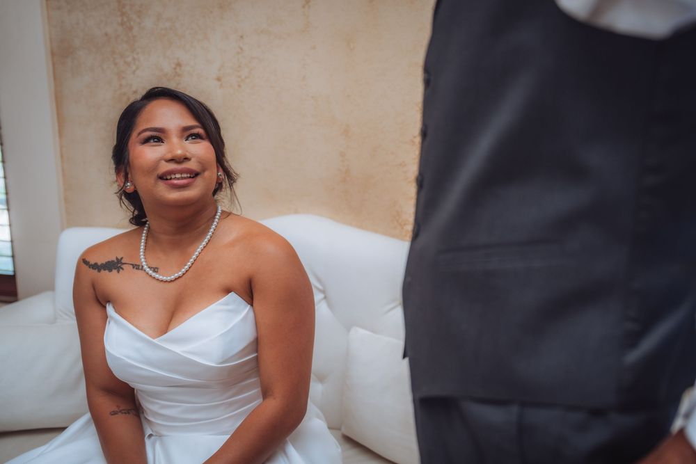 Smiling bride in strapless white gown and pearl necklace sits on white sofa looking up at groom on wedding day.