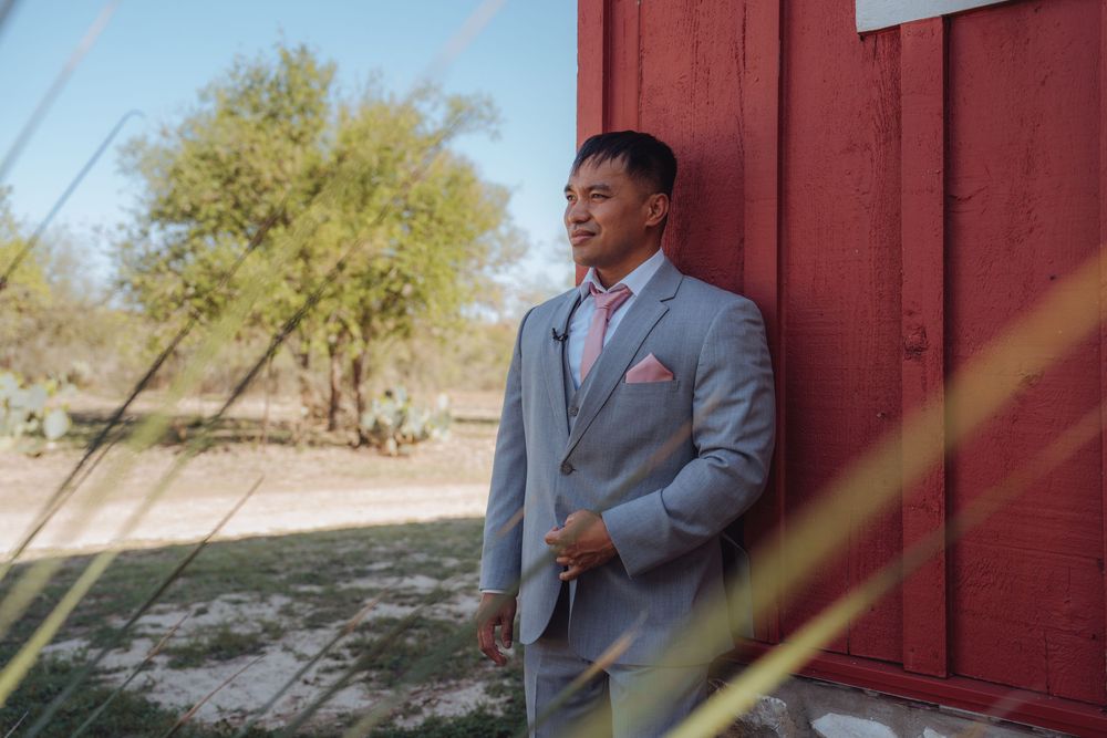 A groom in a light gray suit with pink tie and pocket square leans against a red barn wall outdoors.