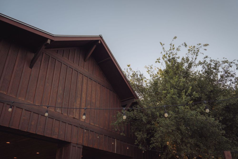 Rustic wooden barn wedding venue exterior with string lights and green trees at dusk.