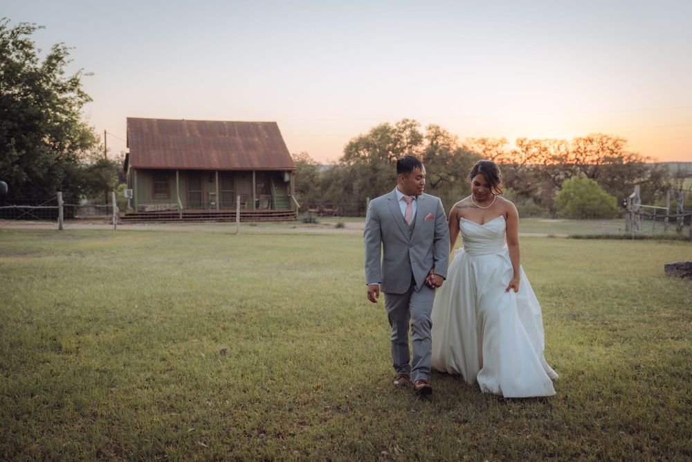 Newlyweds walking hand in hand across a green field at sunset with a rustic barn in the background.