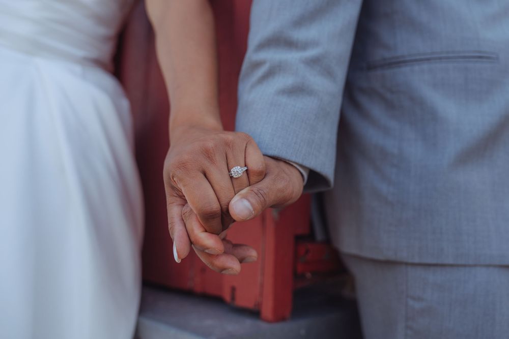 Close-up of newlyweds holding hands, showcasing a diamond engagement ring, with bride in white and groom in gray suit.