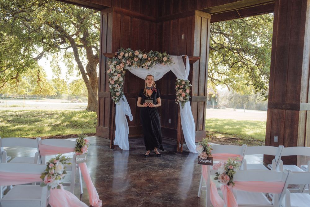 Woman officiant stands beneath floral arch with white draping at rustic outdoor wedding ceremony venue.