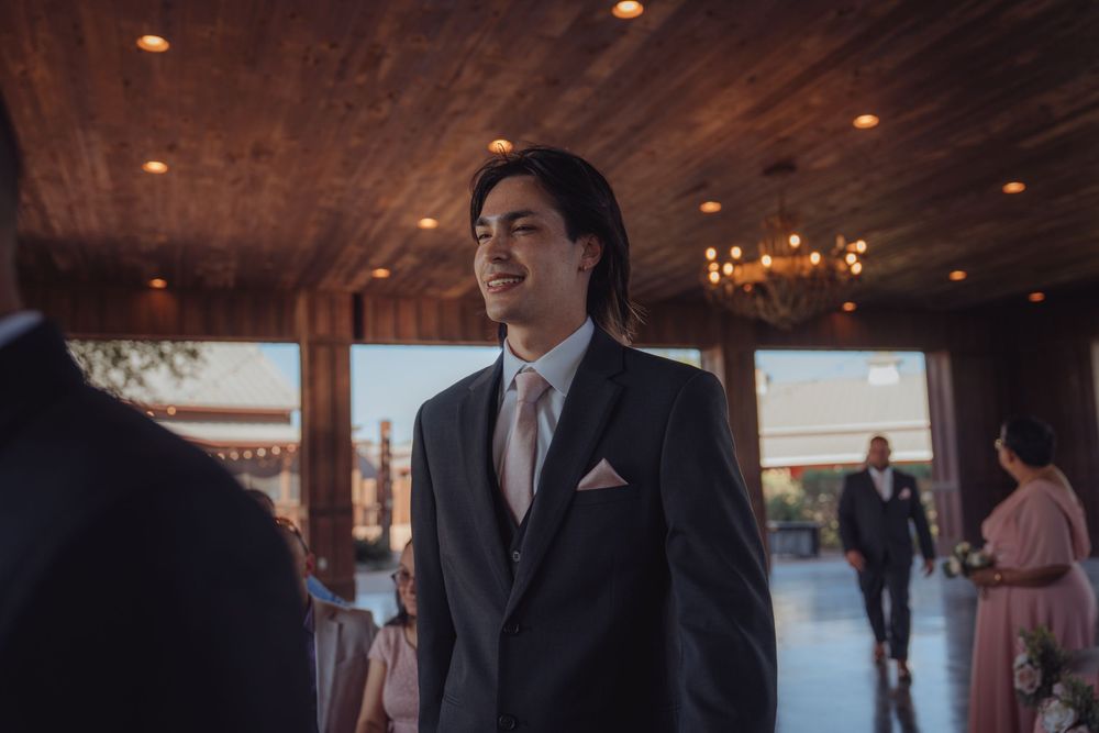 Smiling groom in dark suit with pink tie standing in rustic wedding venue.