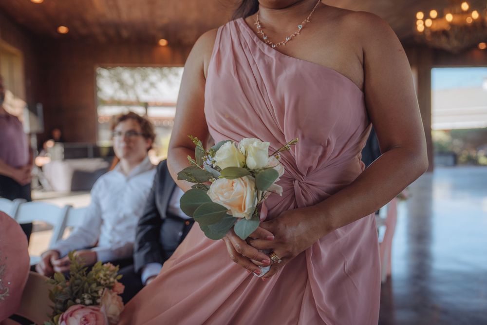 Bridesmaid in blush pink one-shoulder gown holding white and green floral bouquet.