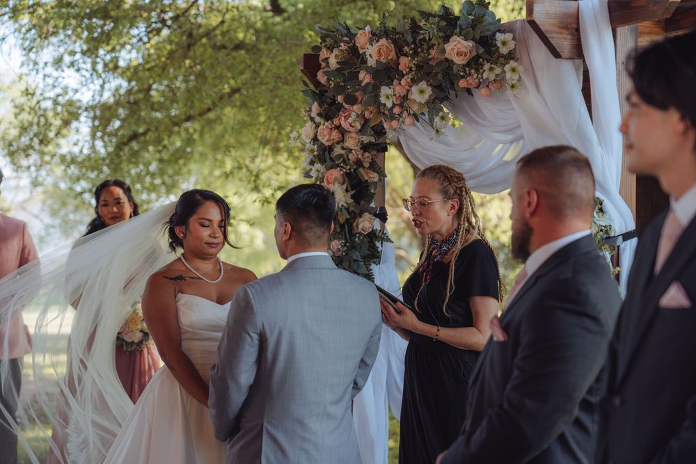 Bride and groom exchange vows outdoors under floral arch with officiant during intimate wedding ceremony.
