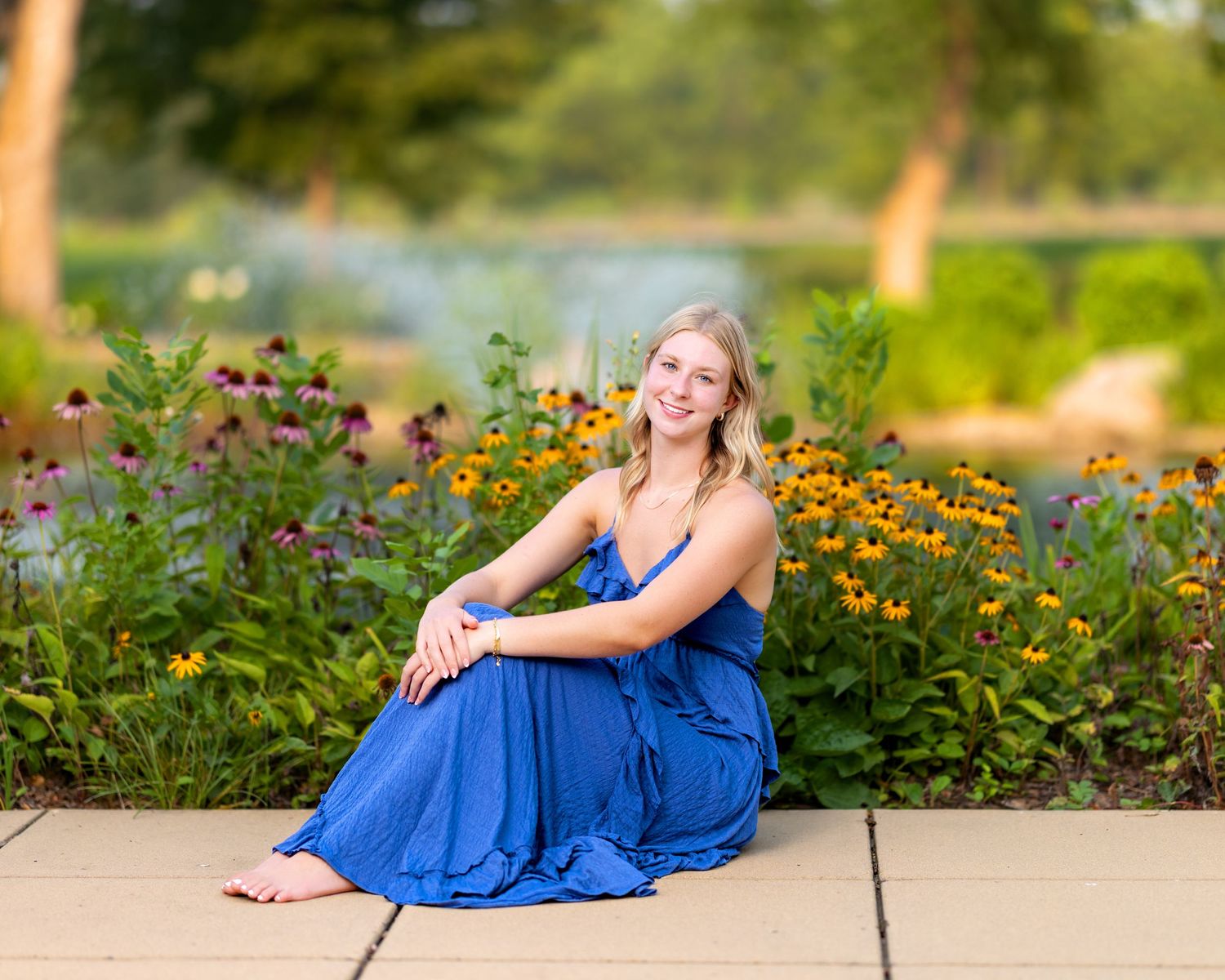 Smiling blonde teen girl in a royal blue maxi dress sits barefoot on a path surrounded by colorful garden flowers.