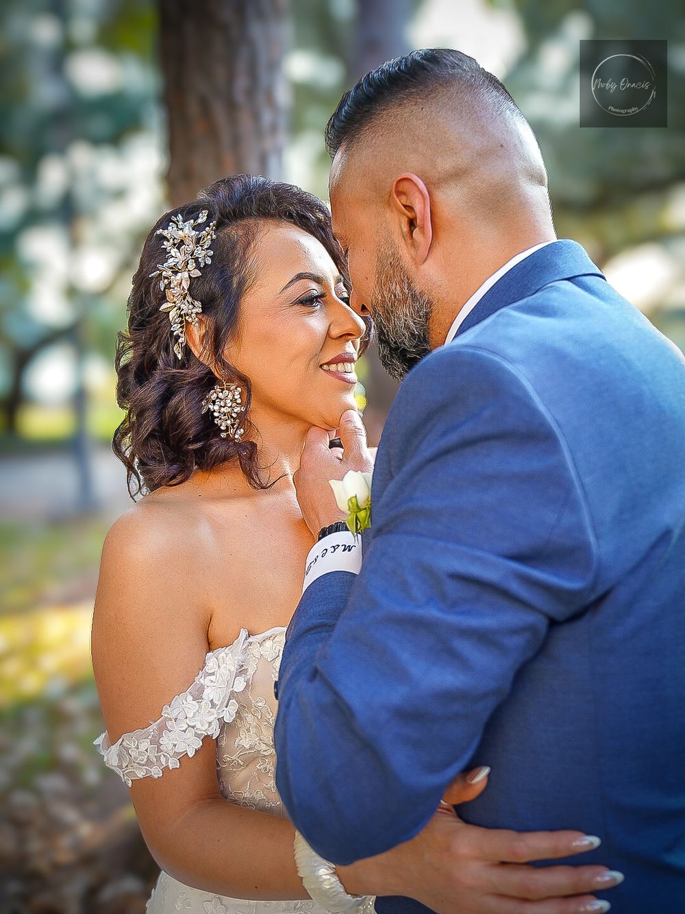Bride and groom share a romantic moment outdoors, with the bride smiling in a lace off-shoulder gown and floral hair accessory.