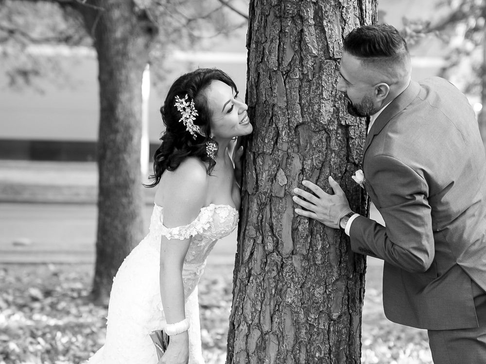 Black and white photo of a bride and groom sharing a romantic moment by a tree, bride wearing a lace gown and floral hairpiece.