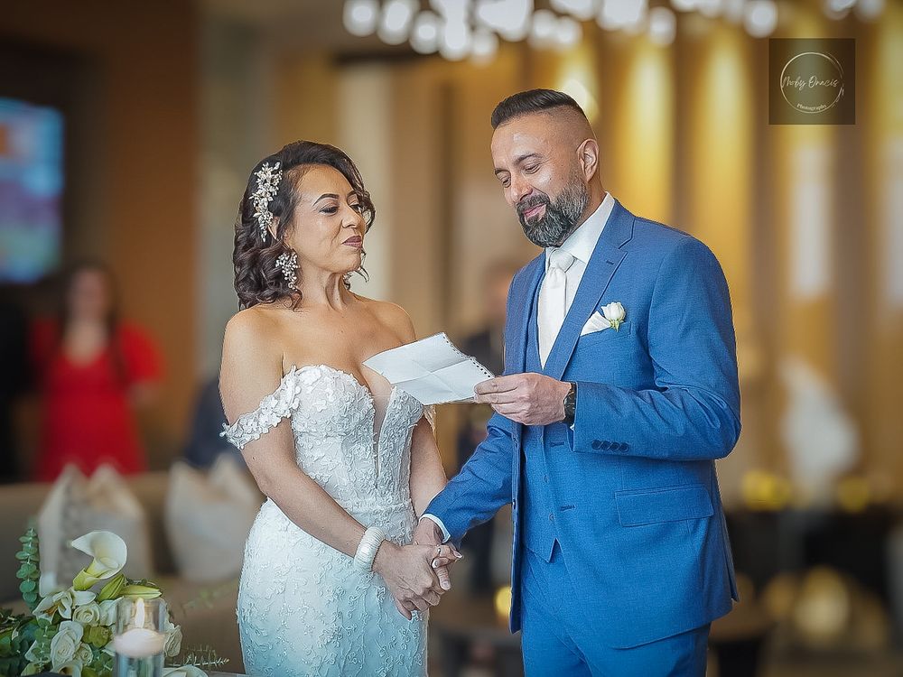 Bride and groom exchange vows during indoor wedding ceremony, groom in blue suit reads from paper.