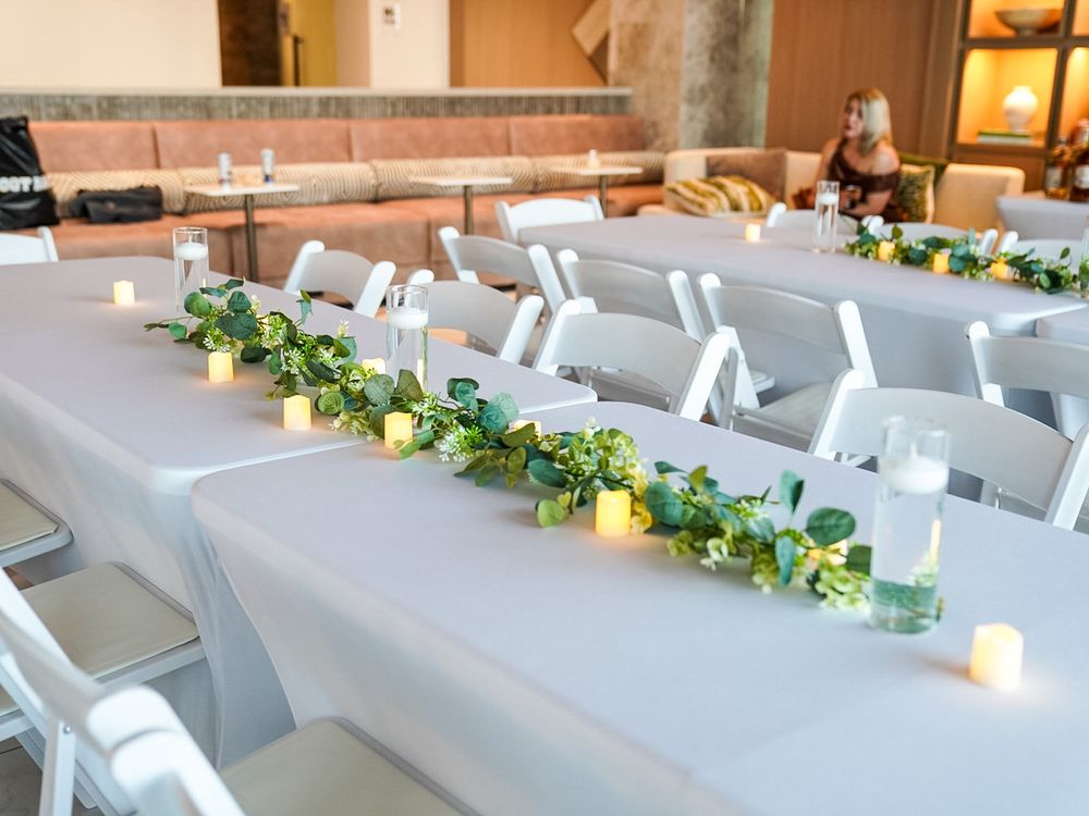 Elegant wedding reception table with white chairs, greenery garland, and glowing candles on gray linen.