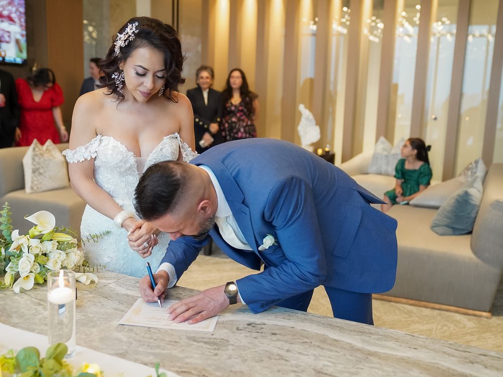 Groom in blue suit signs wedding certificate at marble table while bride in lace gown watches.