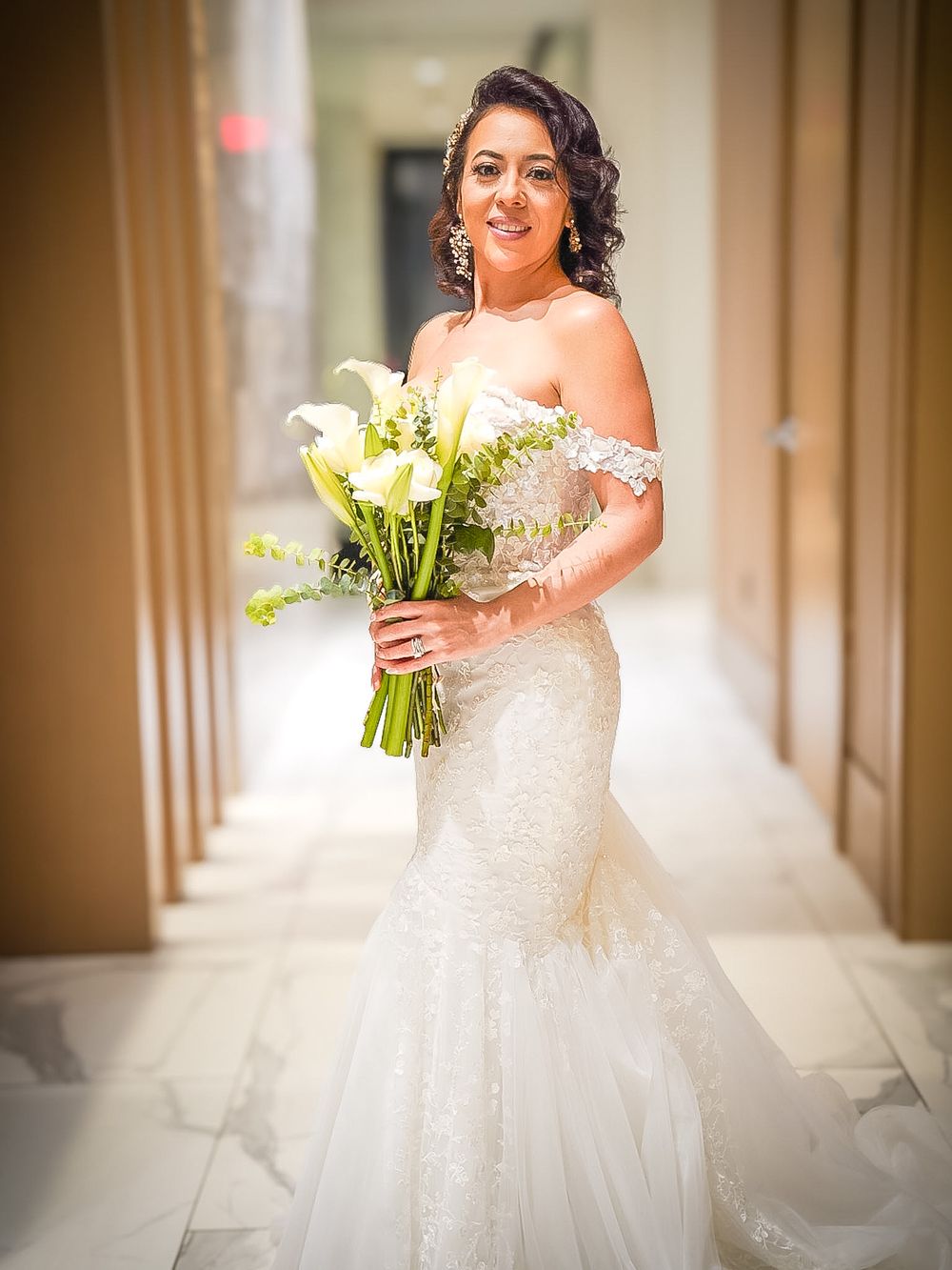 Radiant bride in lace mermaid gown holding white calla lily bouquet in elegant marble hallway.