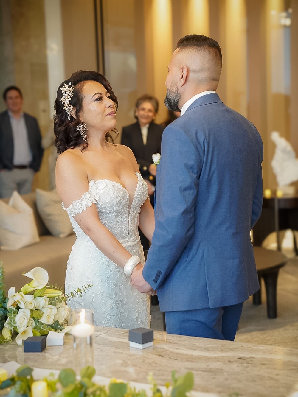 Bride and groom hold hands during intimate indoor wedding ceremony with candles and floral decorations.