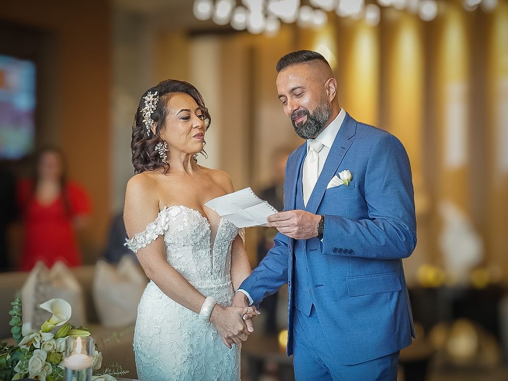 Groom in blue suit reads vows to bride in lace off-shoulder gown during wedding ceremony.