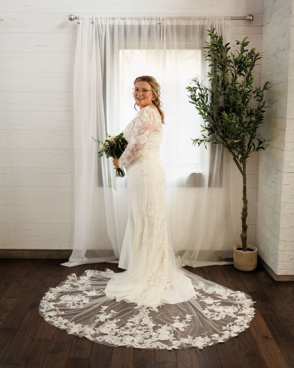 Bride in lace wedding gown with cathedral train holds bouquet, posing by sheer curtains with olive tree backdrop.