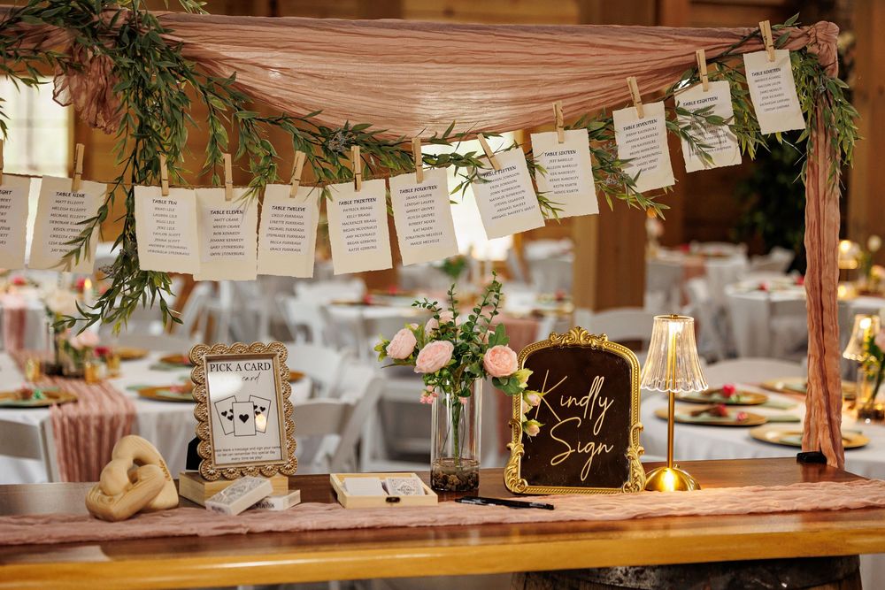 Elegant wedding seating chart display with cards hanging from greenery garland over a rustic wooden table with pink floral decor.