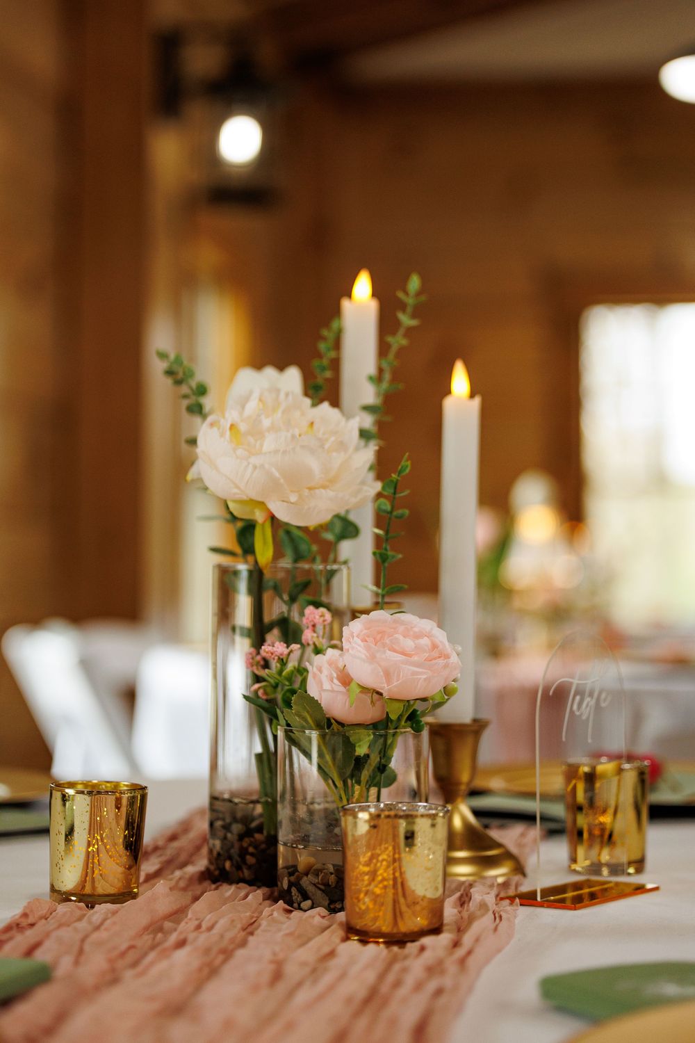 Elegant wedding centerpiece with white peonies, pink roses, gold votives, and white taper candles on a blush table runner.