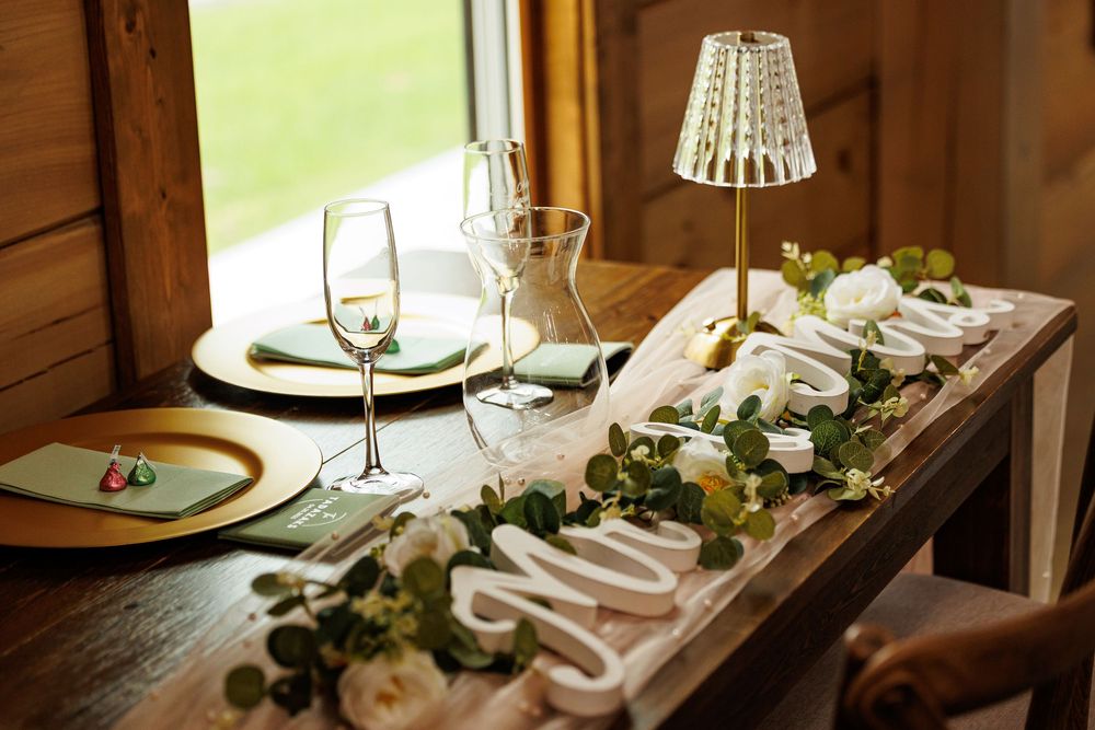 Elegant wedding table setting with champagne glasses, gold accents, white flowers, and greenery garland on rustic wood.