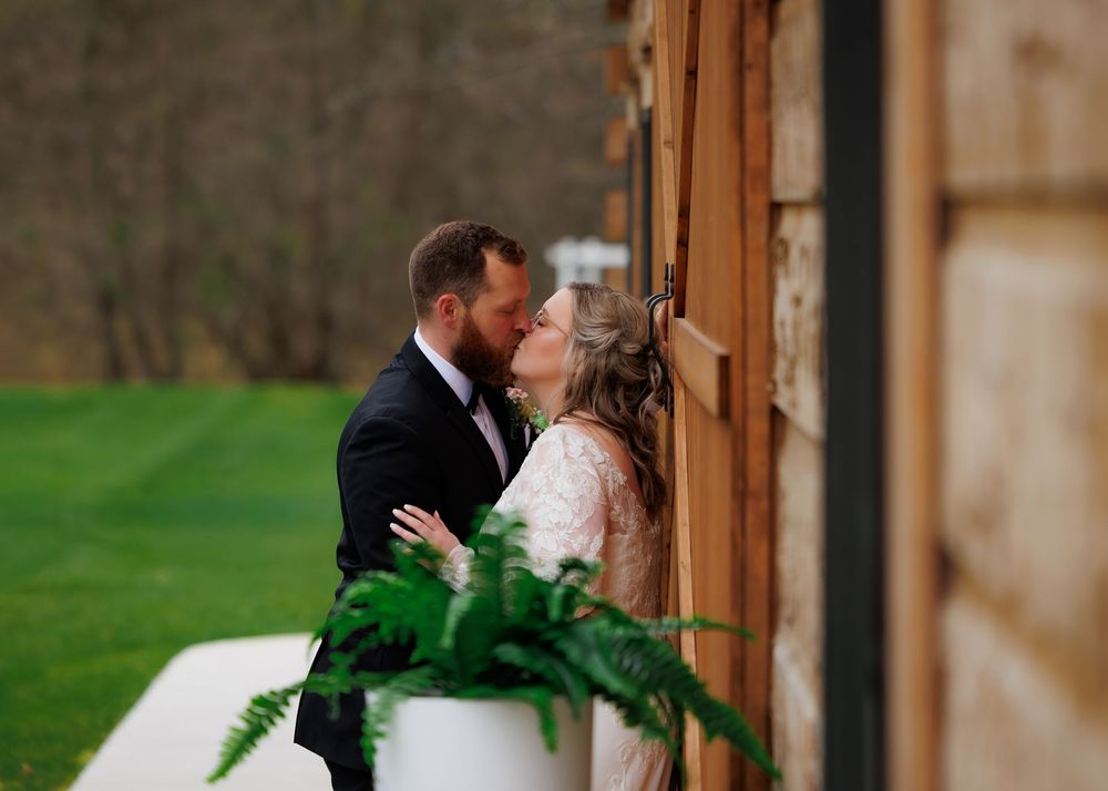 A bride and groom share a romantic kiss beside a rustic wooden barn surrounded by lush green landscape.